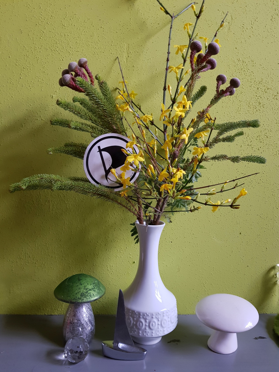 Ein weihnachtlicher Strauss mit Tannenzweigen in einer Vase. Auf den Zweigen liegt ein Aufkleber mit dem Piratenpartei-Logo